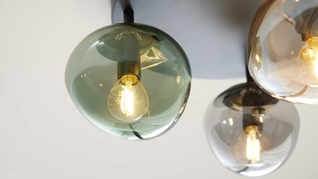 Eine grün getönte hängende Glaskugelleuchte vorn links mit sichtbarem Leuchtfaden, rechts zwei bernstein- und rauchglas-Kugeln.