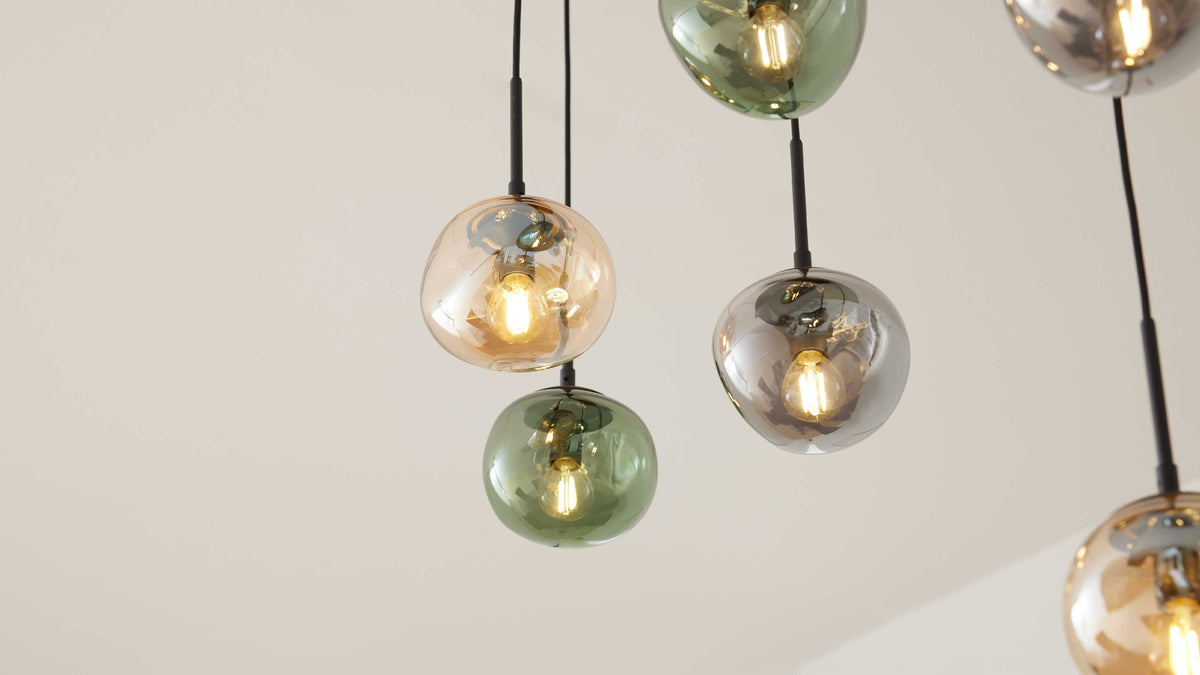 Mehrere hängende Glas-Pendelleuchten mit sichtbaren Filamentlampen und kugeligen Schirmen in Bernstein-, Grün- und Rauchgrau.