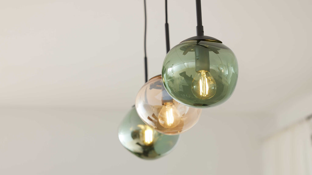 Drei hängende Glaskugelleuchten, vorn ein grüner Schirm mit sichtbarer Filamentlampe, dahinter eine bernstein- und eine weitere grüne Kugel.