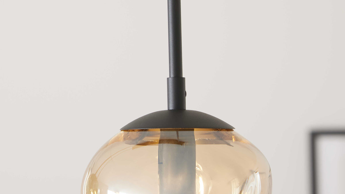 Hängende Pendelleuchte mit bernsteinfarbenem, leicht gewölbtem Glasballon, sichtbarer Leuchtbirne und schwarzem Metallstab mit runder Kappe.