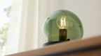 Grüne Glaskugel-Lampe mit sichtbarer, leuchtender Filament-Glühbirne und dunklem Sockel auf Holzfläche.