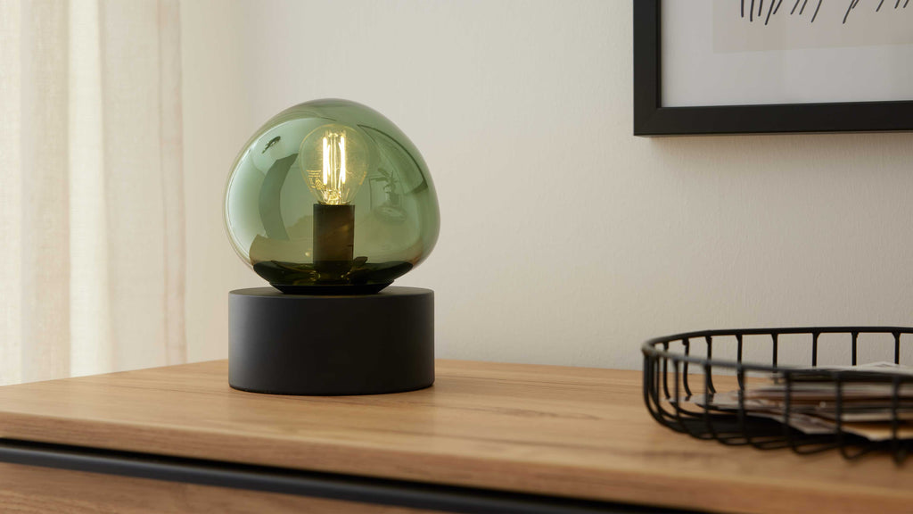 Tischlampe mit grünem Glas-Akzent und schwarzem zylindrischem Sockel steht auf einer Holzkommode, daneben schwarzer Drahtkorb mit Papieren.