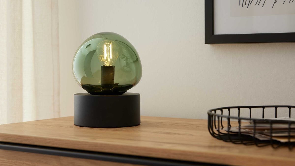 Tischlampe mit grünem Glas-Akzent und schwarzem zylindrischem Sockel steht auf einer Holzkommode, daneben schwarzer Drahtkorb mit Papieren.