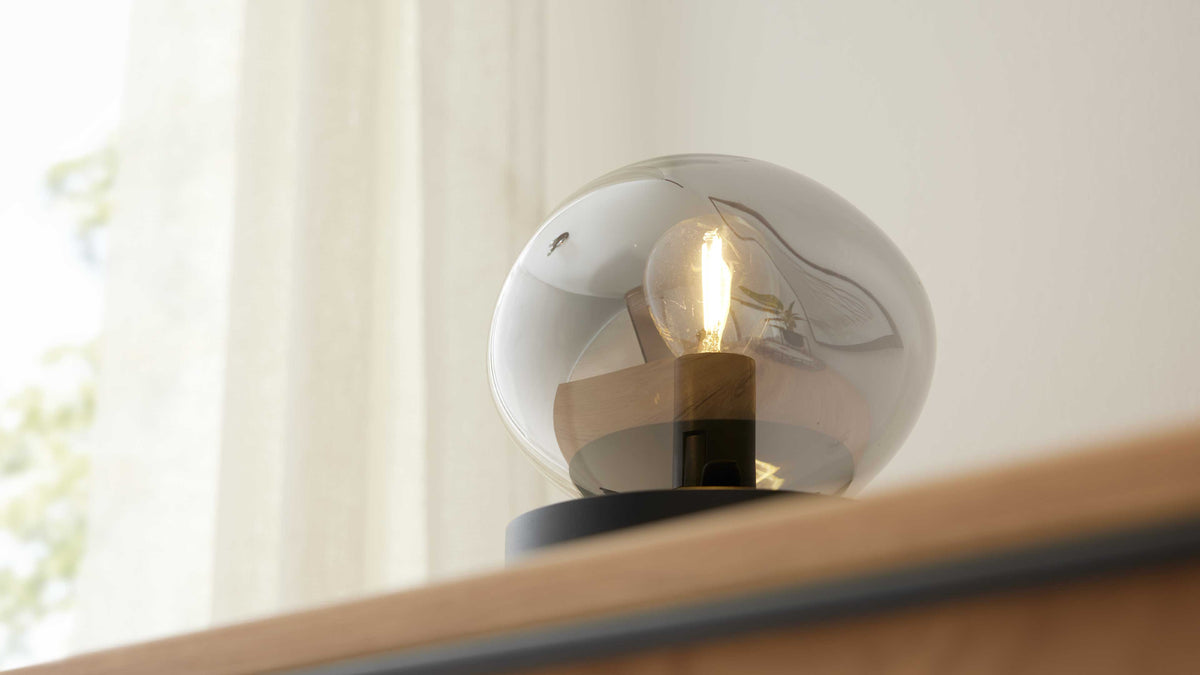 Tischlampe mit rundem rauchfarbenem Glasschirm, sichtbarer Glühbirne, Holzfassung und schwarzem Sockel auf Holzkante