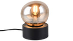 Tischlampe mit runder bernsteinfarbener Glaskugel, sichtbarer Leuchtfadenbirne und schwarzem zylindrischem Sockel mit Kabel.