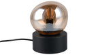 Tischlampe mit rundem schwarzen Zylinderfuß, darauf eine bernsteinfarbene Glasglocke mit sichtbarer Filamentbirne und schwarzem Kabel.