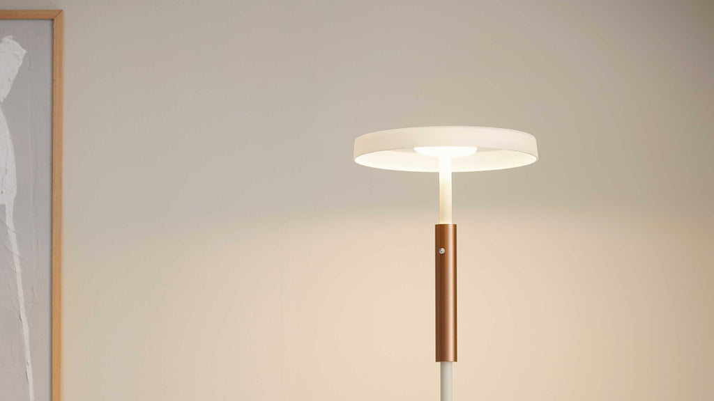 Standlampe mit flachem weißen Rundschirm, schmalem Stab und kupferfarbenem Zylinderdetail, links teilweiser Holzrahmen an der Wand.
