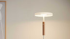 Standlampe mit flachem weißen Rundschirm, schmalem Stab und kupferfarbenem Zylinderdetail, links teilweiser Holzrahmen an der Wand.