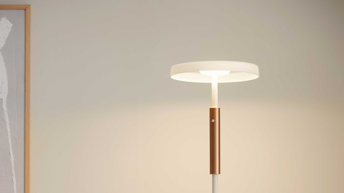 Standlampe mit flachem weißen Rundschirm, schmalem Stab und kupferfarbenem Zylinderdetail, links teilweiser Holzrahmen an der Wand.
