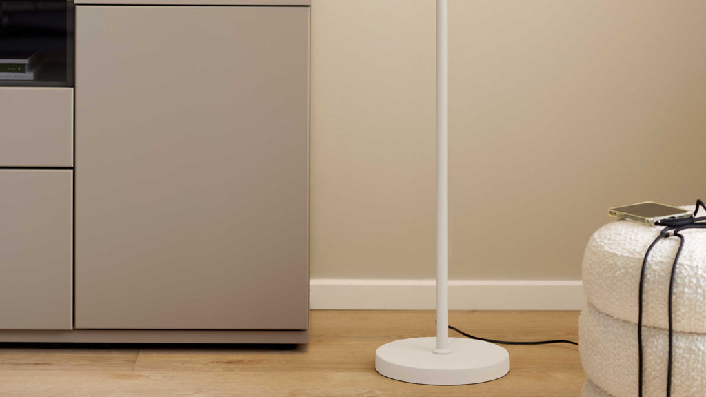 Beige Schrank mit Schubladen links, weiße Stehlampe mit rundem Fuß mittig auf Holzboden und rechts ein Bouclé-Pouf mit Handy und Kabel.