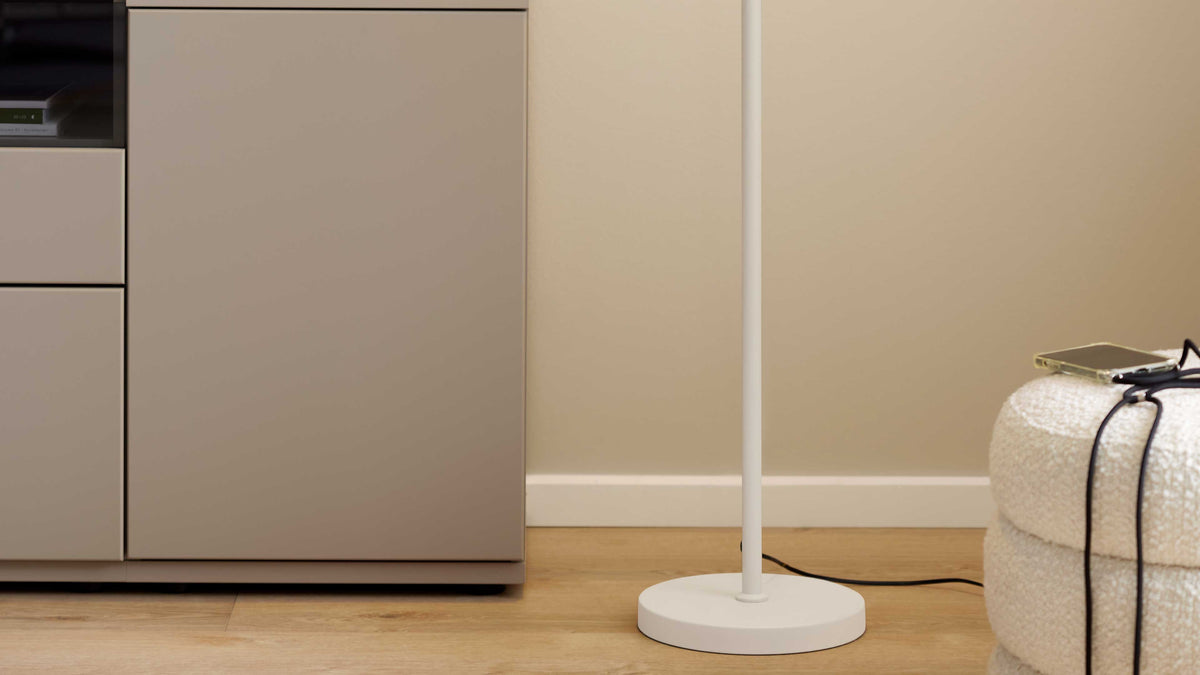 Beige Schrank mit Schubladen links, weiße Stehlampe mit rundem Fuß mittig auf Holzboden und rechts ein Bouclé-Pouf mit Handy und Kabel.