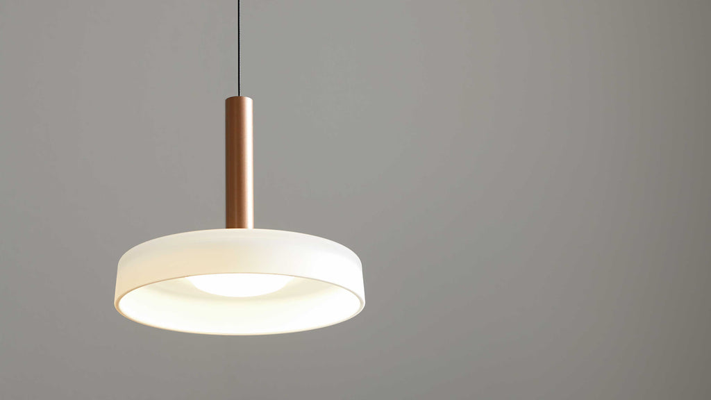 Hängende Deckenlampe mit weißem ringförmigem Schirm, zentraler Lichtquelle und kupferfarbenem Zylinder sowie schwarzem Kabel vor grauer Wand.