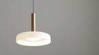 Hängende Deckenlampe mit weißem ringförmigem Schirm, zentraler Lichtquelle und kupferfarbenem Zylinder sowie schwarzem Kabel vor grauer Wand.