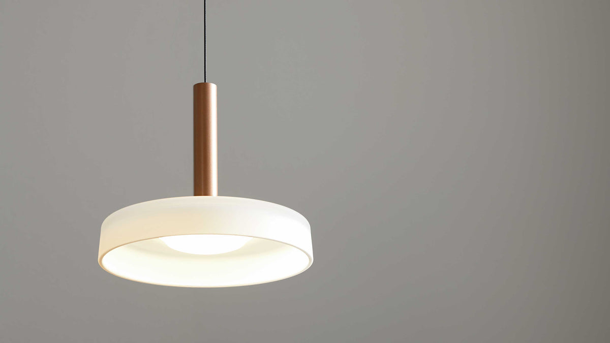 Hängende Deckenlampe mit weißem ringförmigem Schirm, zentraler Lichtquelle und kupferfarbenem Zylinder sowie schwarzem Kabel vor grauer Wand.