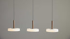 Drei hängende Deckenlampen mit braunen zylindrischen Stangen und flachen weißen Diffusoren, an schwarzen Kabeln vor grauer Wand.