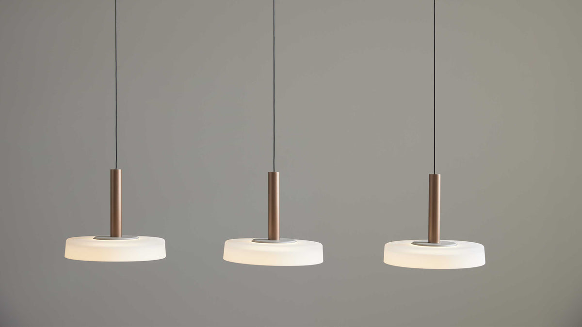 Drei hängende Deckenlampen mit braunen zylindrischen Stangen und flachen weißen Diffusoren, an schwarzen Kabeln vor grauer Wand.