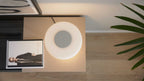 Rundliche, ringförmige Tischlampe mit warmem Licht auf rechteckiger beige Ablage neben Magazin und Pflanzenblatt.