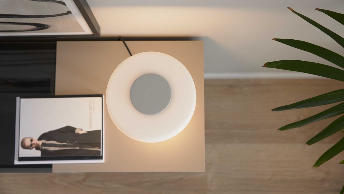 Rundliche, ringförmige Tischlampe mit warmem Licht auf rechteckiger beige Ablage neben Magazin und Pflanzenblatt.