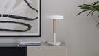 Tischlampe mit weißem Rundschirm und kupferfarbenem Schaft auf beigem Sideboard; links schwarz gerahmtes Bild, Magazin und Pflanzblätter rechts.