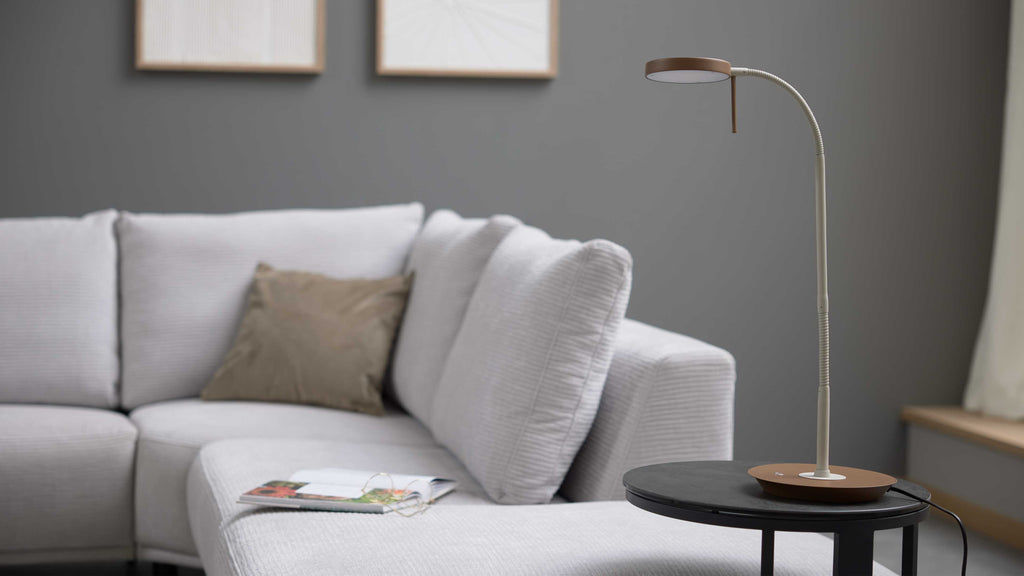 Stehlampe mit braunem Sockel auf rundem schwarzen Beistelltisch, davor hellgraues Sofa mit Kissen und Zeitschrift.