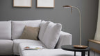 Stehlampe mit braunem Sockel auf rundem schwarzen Beistelltisch, davor hellgraues Sofa mit Kissen und Zeitschrift.