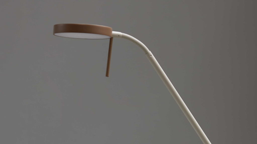 Schreibtischlampe mit rundem braunem Lampenschirm und weißem, gebogenem Schwanenhals sowie langem Stabarm vor grauem Hintergrund.