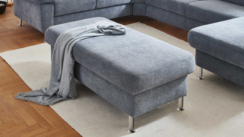Grau gepolsterter Hocker mit Metallfüßen, darauf liegt ein grauer Überwurf, vor einem hellen Teppich auf Holzfußboden.