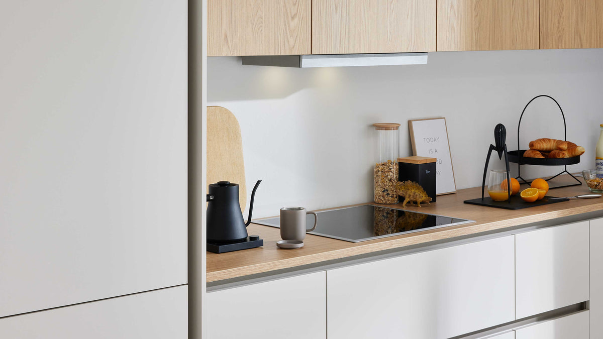 Küchenarbeitsfläche mit hellem Holz, schwarzem Kaffeekännchen, grauer Tasse, Induktionsfeld, Müsliglas, Obstkorb und Croissants.