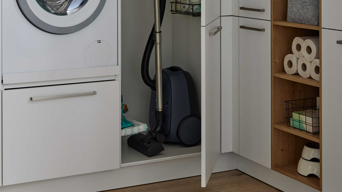 Offener weißer Schrank mit blauem Staubsauger, links Waschmaschinenfront, rechts offene Holzregale mit Toilettenpapier, Drahtkorb und Napf.