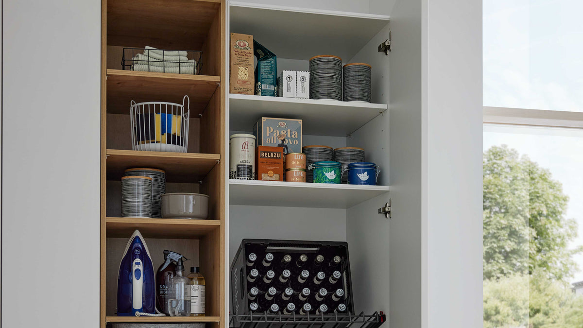 Offener Schrank mit Holzregalen links (Bügeleisen, Flaschen, Schalen, Drahtkorb) und rechten Regalböden mit Dosen, Kartons und Bierkiste.