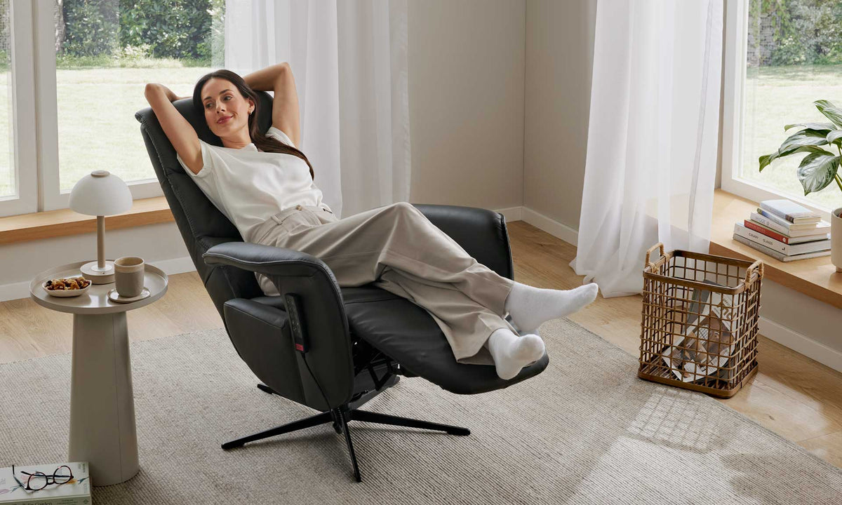 Eine Frau entspannt sich in einem HUKLA® | IL Relaxsessel der Interliving-Serie 4545 aus Glattleder, die Füße hochgelegt, in einem sonnendurchfluteten Wohnzimmer.