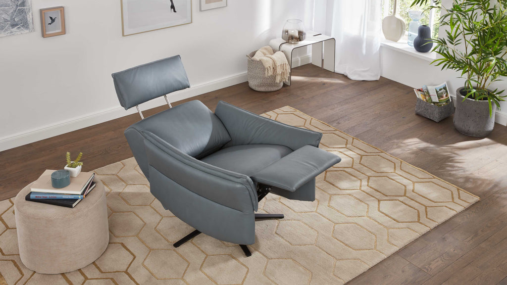 Ein grauer, gepolsterter Sessel mit verstellbarem Rücken, auf einem beigen Teppich mit geometrischem Muster, steht neben einem kleinen Beistelltisch.