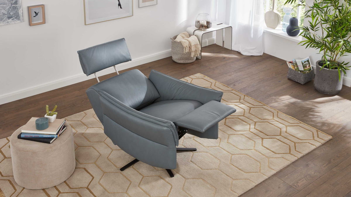 Ein grauer, gepolsterter Sessel mit verstellbarem Rücken, auf einem beigen Teppich mit geometrischem Muster, steht neben einem kleinen Beistelltisch.
