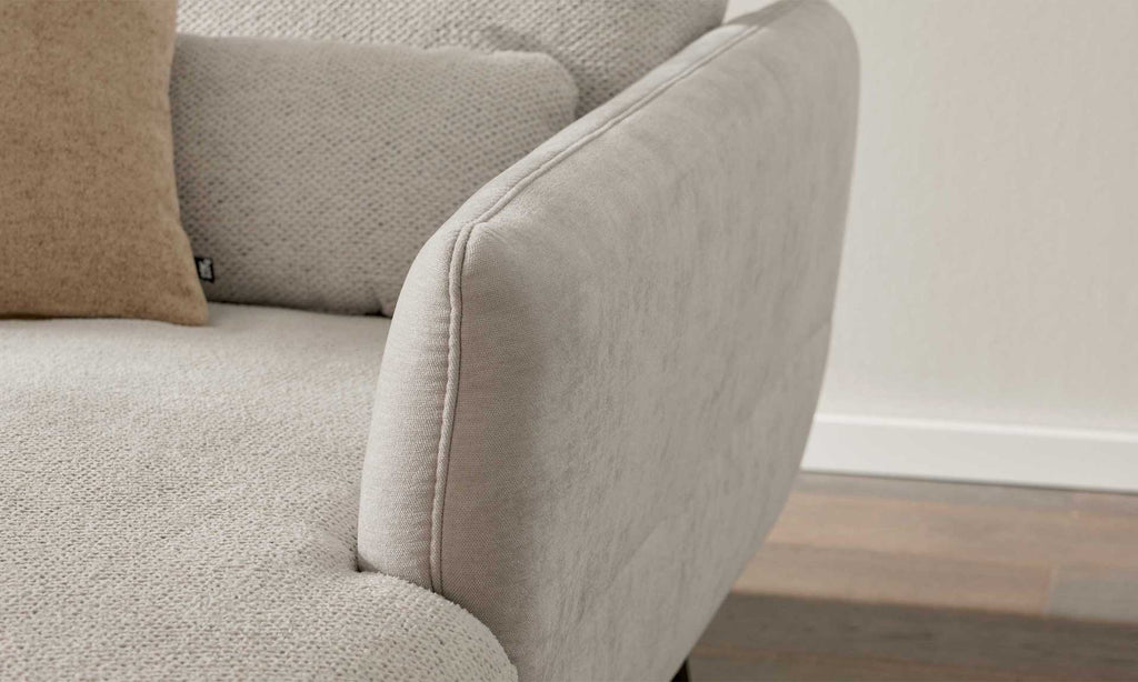 Eine Couch mit hellem, strukturiertem Bezug, einem abgerundeten Armlehnenbereich und einem beigen Kissen im Hintergrund.