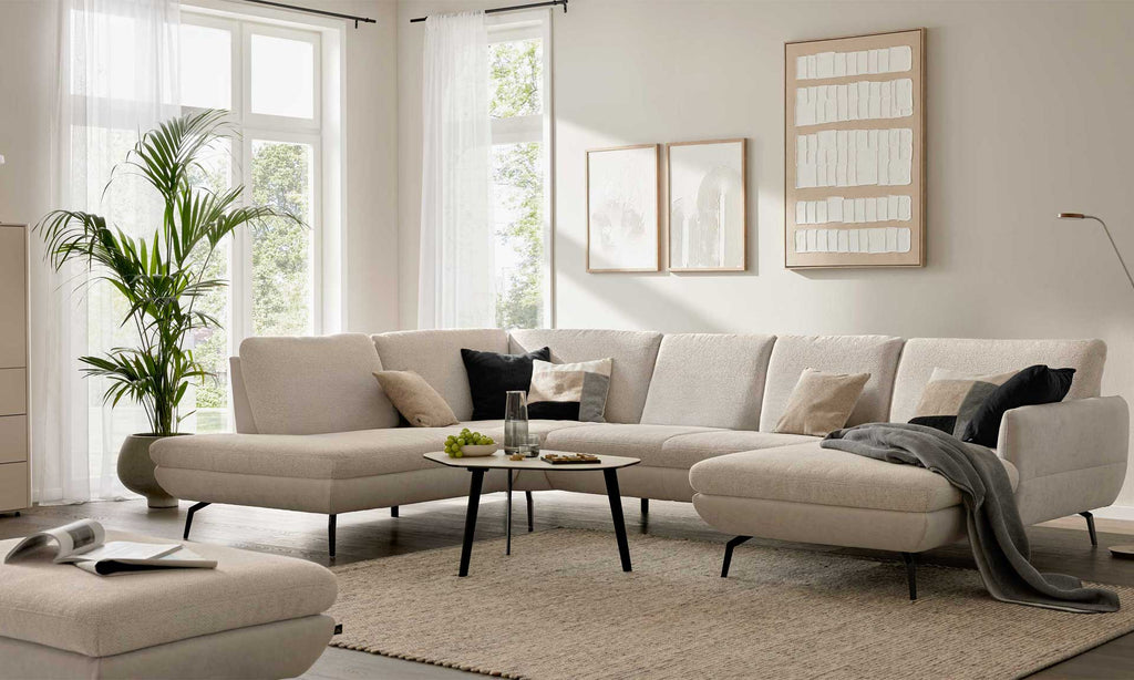Eine helle, geräumige Wohnlandschaft mit hohen Armlehnen, mehreren Kissen, einem kleinen Tisch und einer Pflanze neben großen Fenstern.