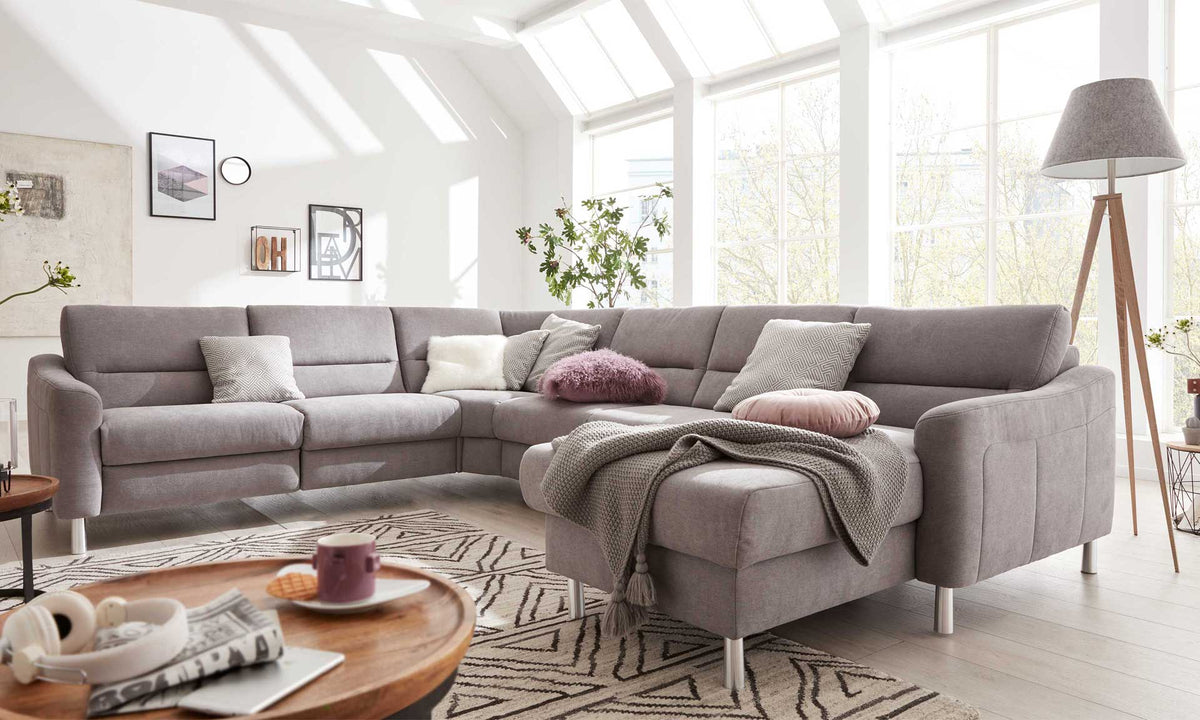 Eine graue L-förmige Couch mit verschiedenen Kissen und einer Decke, umgeben von einem runden Tisch und einer Stehlampe.