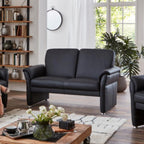 Modernes Wohnzimmer mit einem Sofa „Bornholm II“ von Steinpol Polsteria, einem Holztisch, einer Pflanze, Regalen und Sonnenlicht, das durch große Fenster hereinfällt.