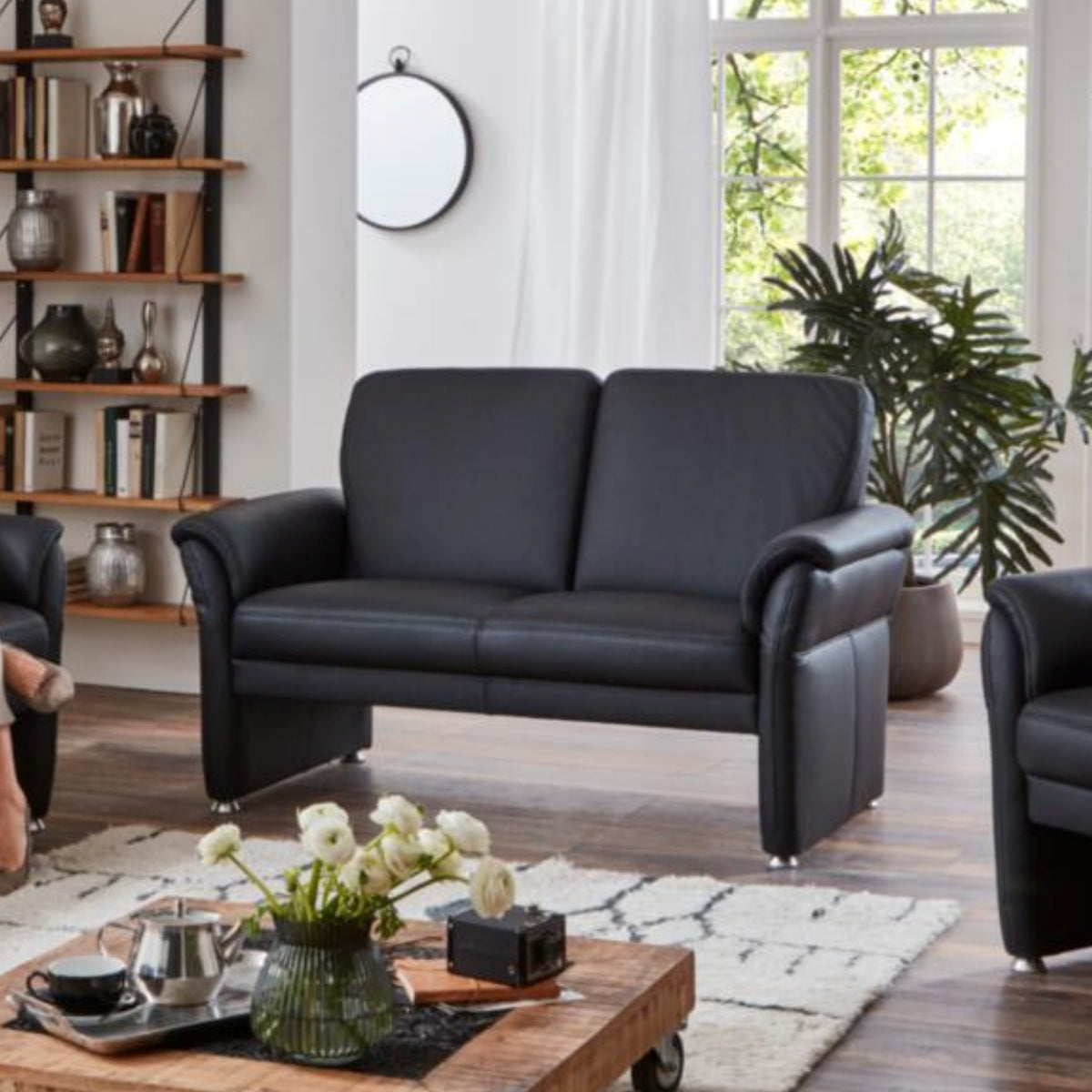 Modernes Wohnzimmer mit einem Sofa „Bornholm II“ von Steinpol Polsteria, einem Holztisch, einer Pflanze, Regalen und Sonnenlicht, das durch große Fenster hereinfällt.