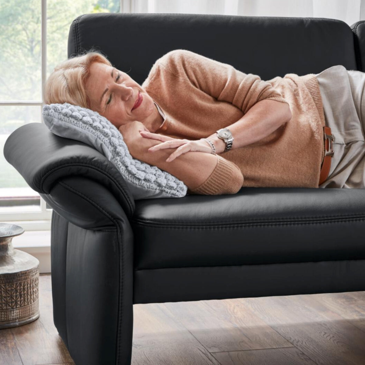Eine lächelnde Frau auf dem Sofa „Steinpol Polsteria Bornholm II“, die sich auf einem grauen Kissen im Sonnenlicht entspannt.