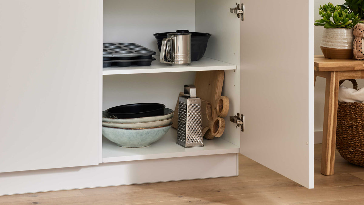 Im offenen Schrank sind übereinandergestapelte Schalen, ein Muffinblech, ein Kännchen, ein Holzbrett und eine Reibe sichtbar.