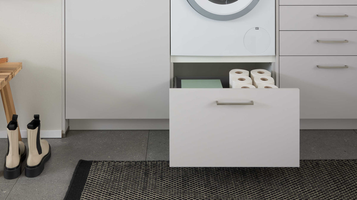 Ein offenes, graues Schubladenmöbel zeigt darin mehrere Rollen Toilettenpapier und einen grünen Ordner, daneben steht ein kleiner Holzregal.