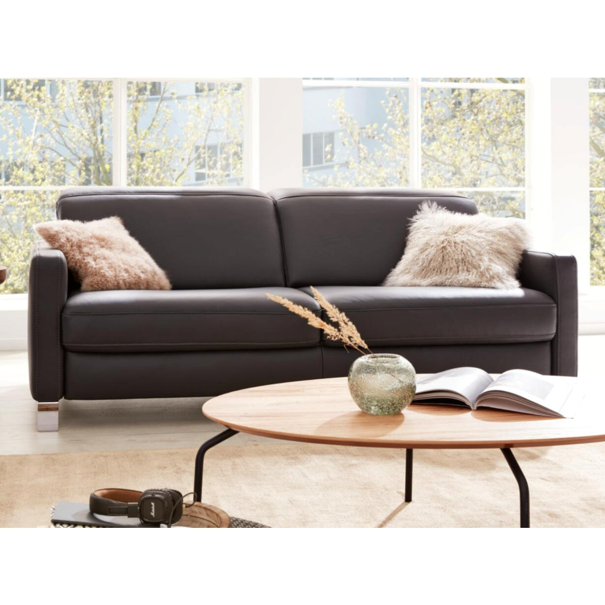 Hinter einem runden Couchtisch steht ein Sofa der HUKLA® IL Interliving Serie 4054 in Leder Anthrazit mit zwei Plüschkissen.