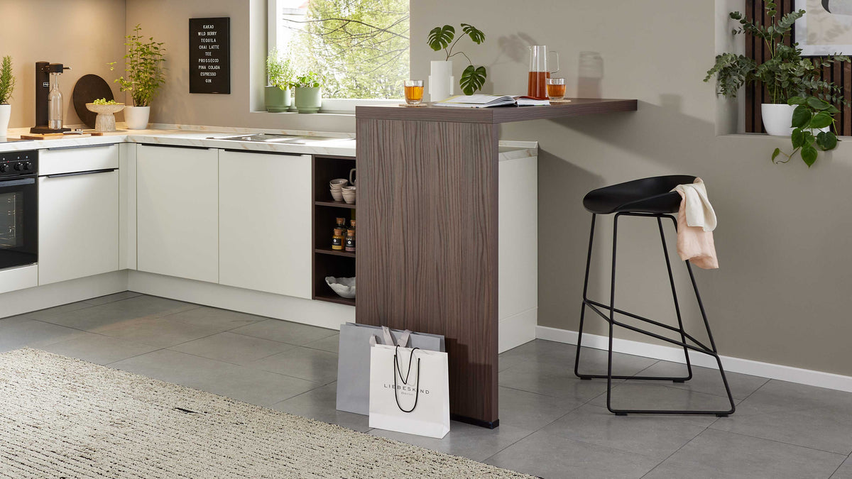 In einer modernen Küche steht eine Holzbar mit einer schwarzen Barhocker, daneben liegen zwei Einkaufstüten auf dem grauen Fliesenboden.