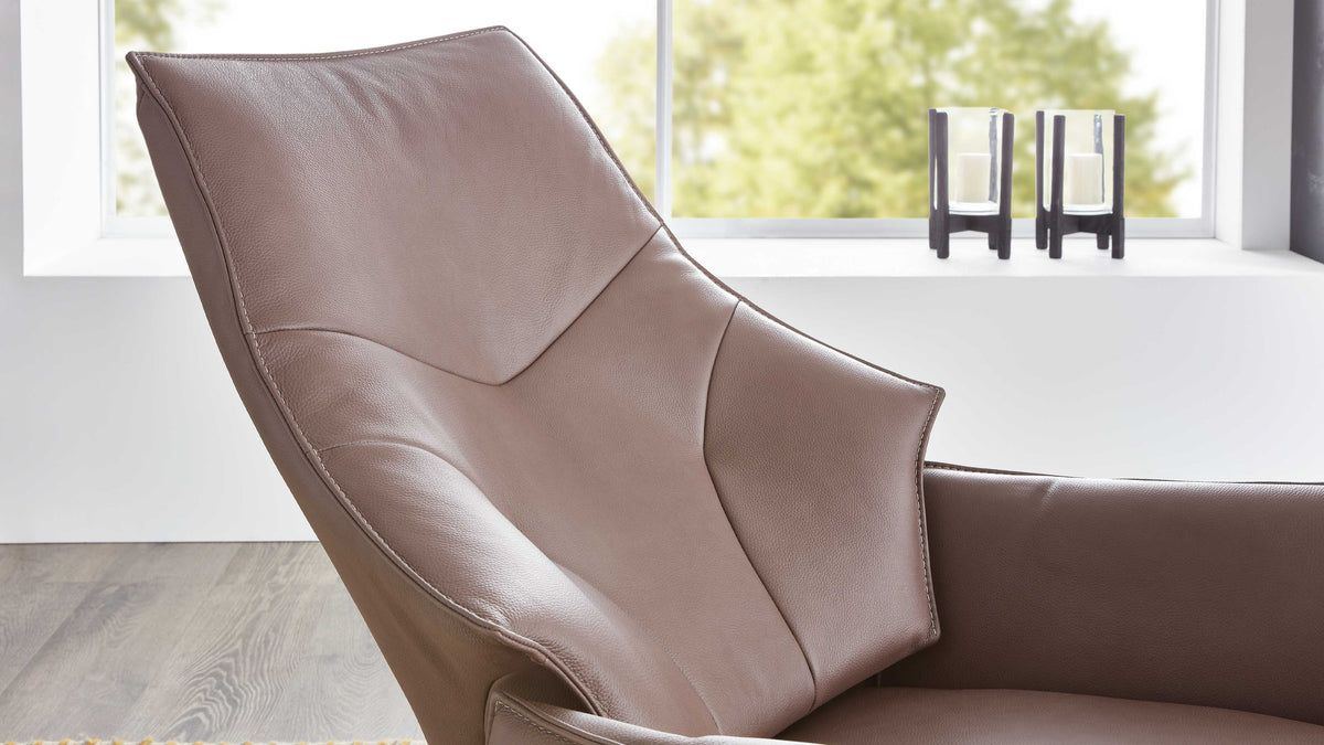 Beige Lounge-Sessel mit hohen, geschwungenen Armlehnen und gepolsterter Rückenlehne vor einem Fenster, neben zwei Kerzenhaltern.