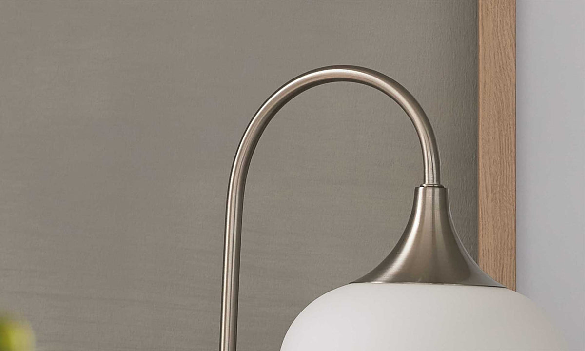 Eine Lampe mit einem gebogenen Metallarm in Stahloptik und einem weißen satinierten Glasschirm vor einer grauen Wand.