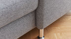 Ecke eines grauen Sofas mit einem textilem Bezug und einem silbernen Metallfuß auf einem Holzboden.