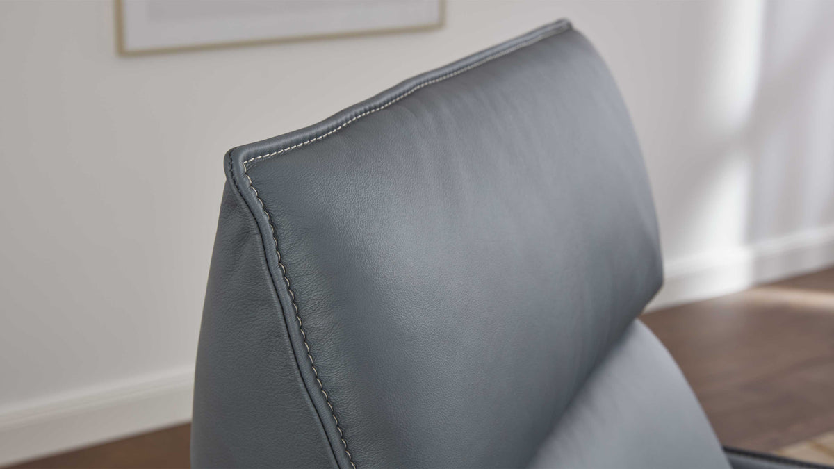Ein grauer Lederstuhl ist im Fokus, der ein schlichtes Design mit einer umschlossenen Rückenlehne und sichtbaren Nähten aufweist.