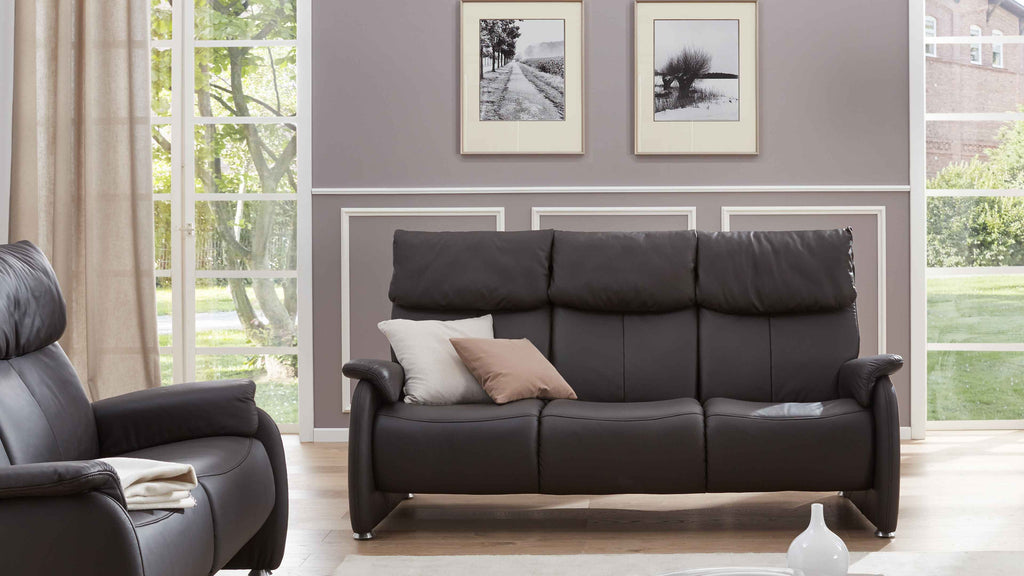 Ein dunkelgraches Sofa mit hohen Rückenlehnen und Kissen steht vor einer Wand mit zwei schwarz-weißen Bildern, daneben ein Fenster mit Blick ins Grüne.