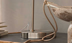 Eine stilvolle Tischlampe mit einem runden, metallischen Fuß steht auf einer dunklen Oberfläche, daneben liegt ein grauer Steinbehälter und Bücher.