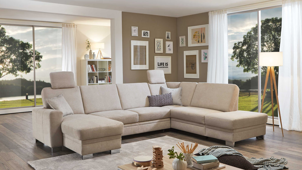 Eine helle, U-förmige Couch mit erhöhtem Kopfteil und einem kleinen Kissen steht auf einem hellen Teppich in einem Wohnzimmer mit großen Fenstern.